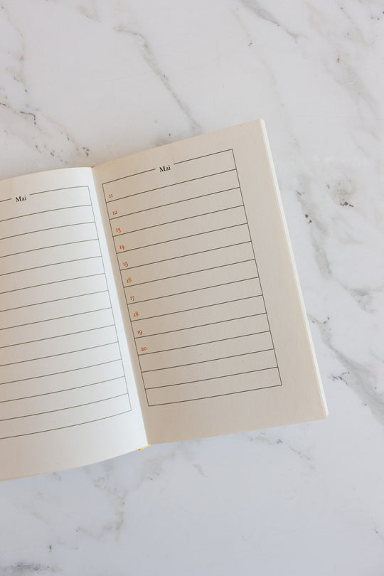 A book lies open on a white marble surface, showing a double-page layout with lined sections and red dates in German for May. The mostly blank pages highlight the spacious area for notes and planning.