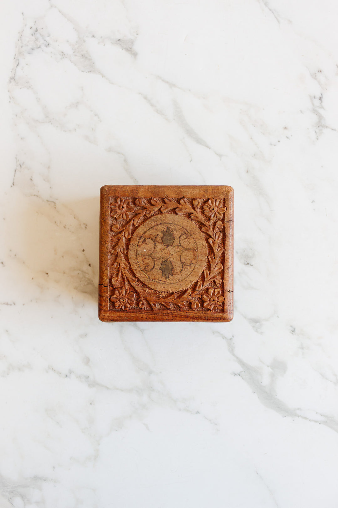 The wood box is small and square, featuring ornate floral carvings on its lid. Resting on a white marble surface, its compact size makes it ideal for storing keepsakes or jewelry.