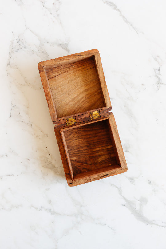 The wood box, small and empty with a hinged lid, sits open to show its smooth interior and compact size on a white marble surface with light gray veining.