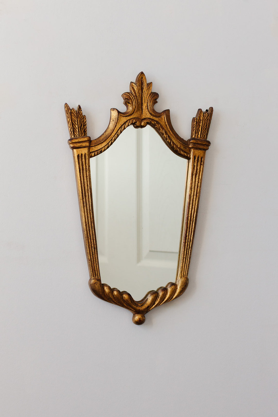 A large ornate gold-framed mirror with intricate detailing hangs on a plain white wall, reflecting a white paneled door.