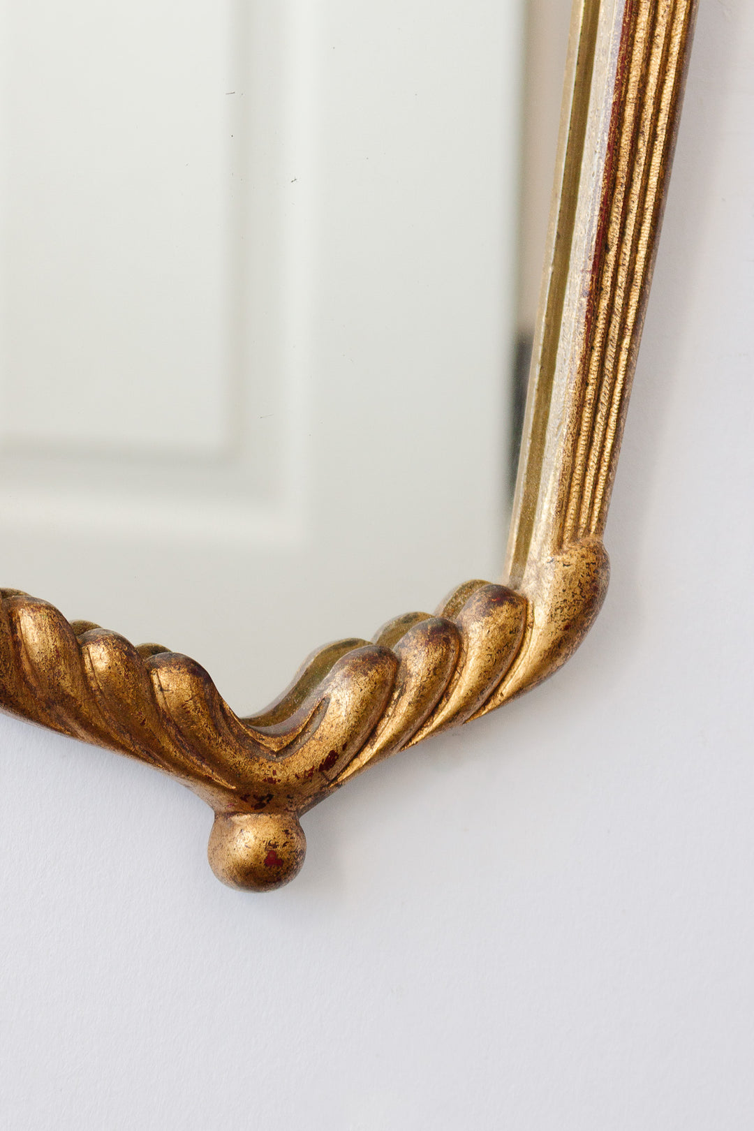 Close-up of the ornate gold corner of the mirror against a white wall, capturing its elegant decorative frame details and a subtle reflection of a nearby door.