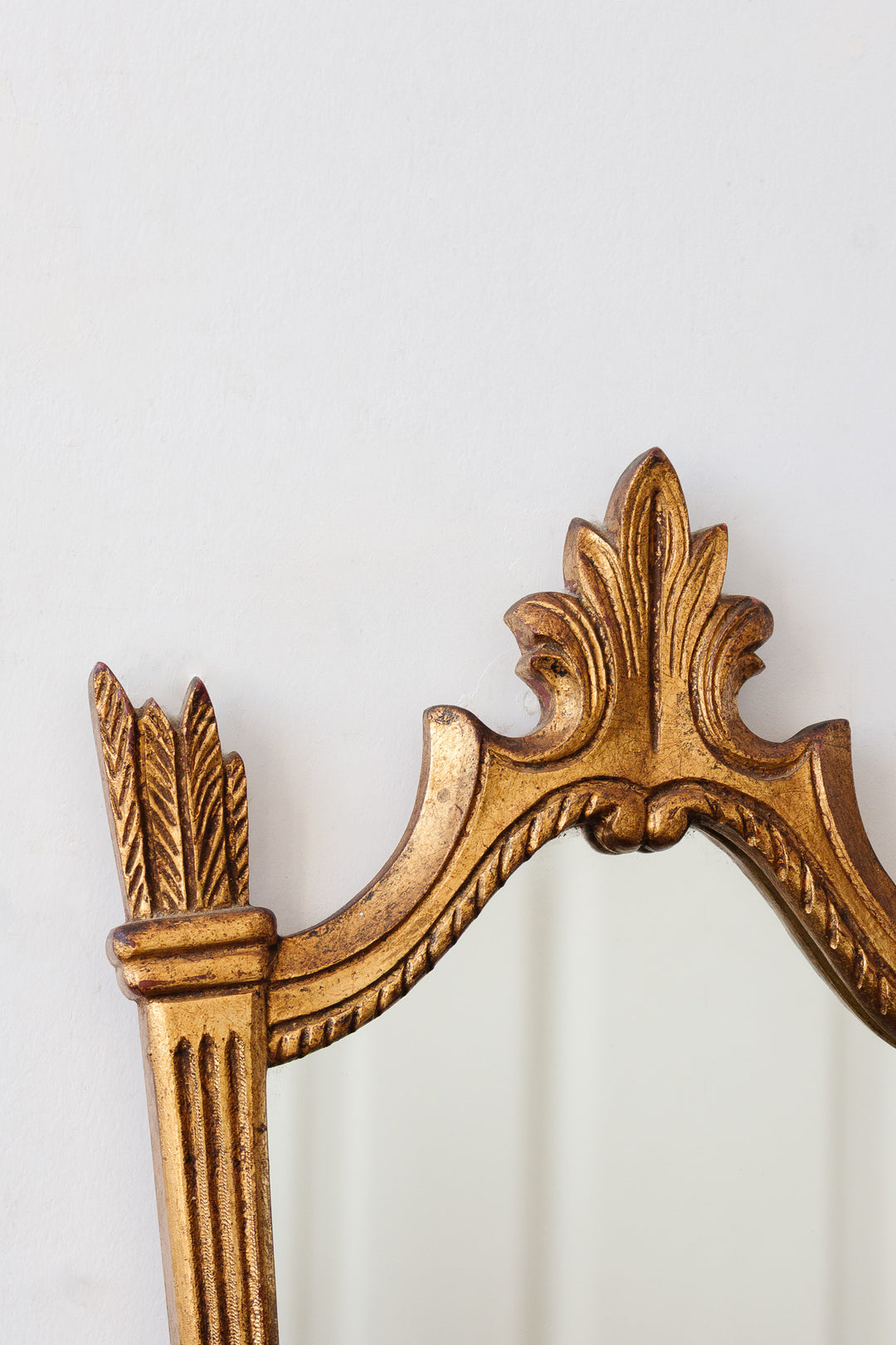 Close-up of the ornate top corner of a gold-framed mirror, showing decorative leaf and column details that emphasize its elegant look against a plain white wall.