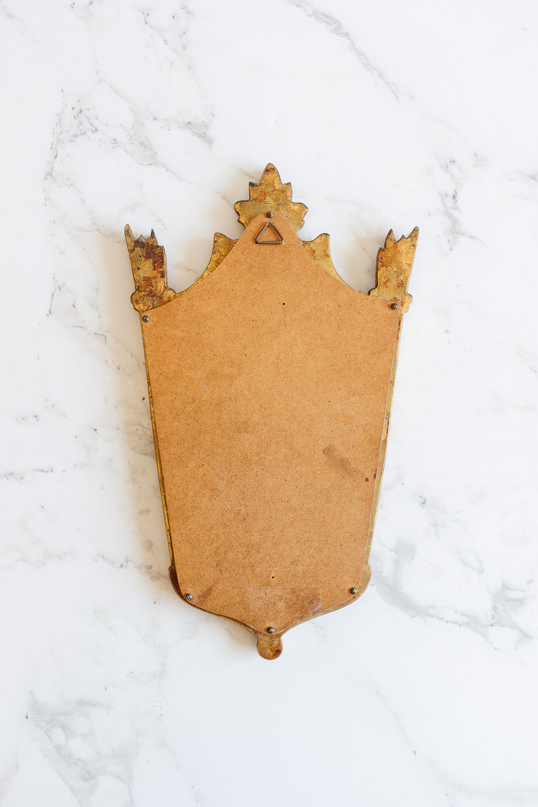 A shield-shaped mirror with ornate wooden edges and a plain back sits on a white marble surface. Showing slight wear and corner metal fasteners, this decorative mirror's versatile size makes it ideal for display.