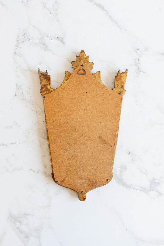 A shield-shaped mirror with ornate wooden edges and a plain back sits on a white marble surface. Showing slight wear and corner metal fasteners, this decorative mirror's versatile size makes it ideal for display.