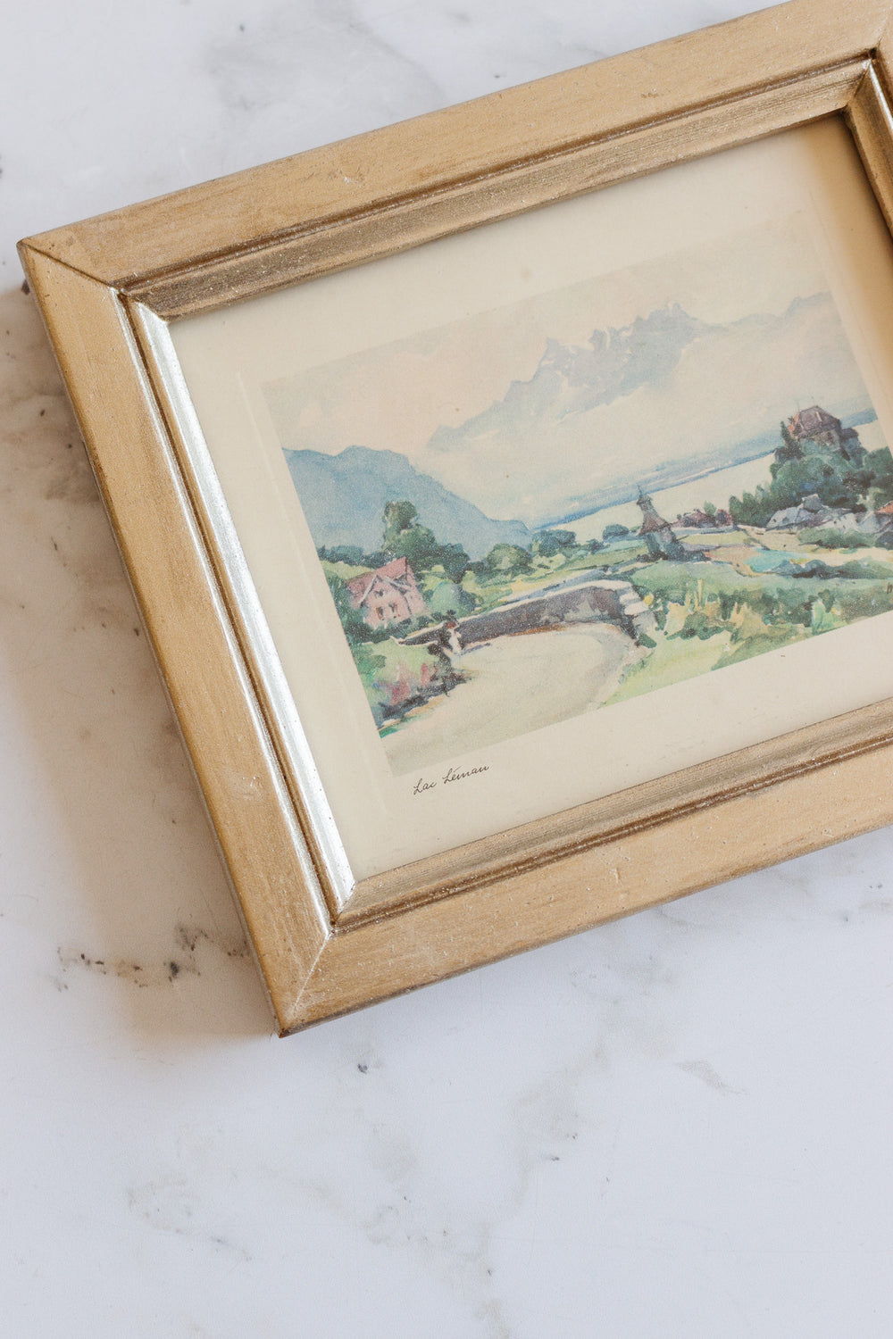 The art is a framed watercolor landscape with mountains, trees, and a small bridge, displayed on a white marble surface. Its dimensions make it an ideal accent for any space.