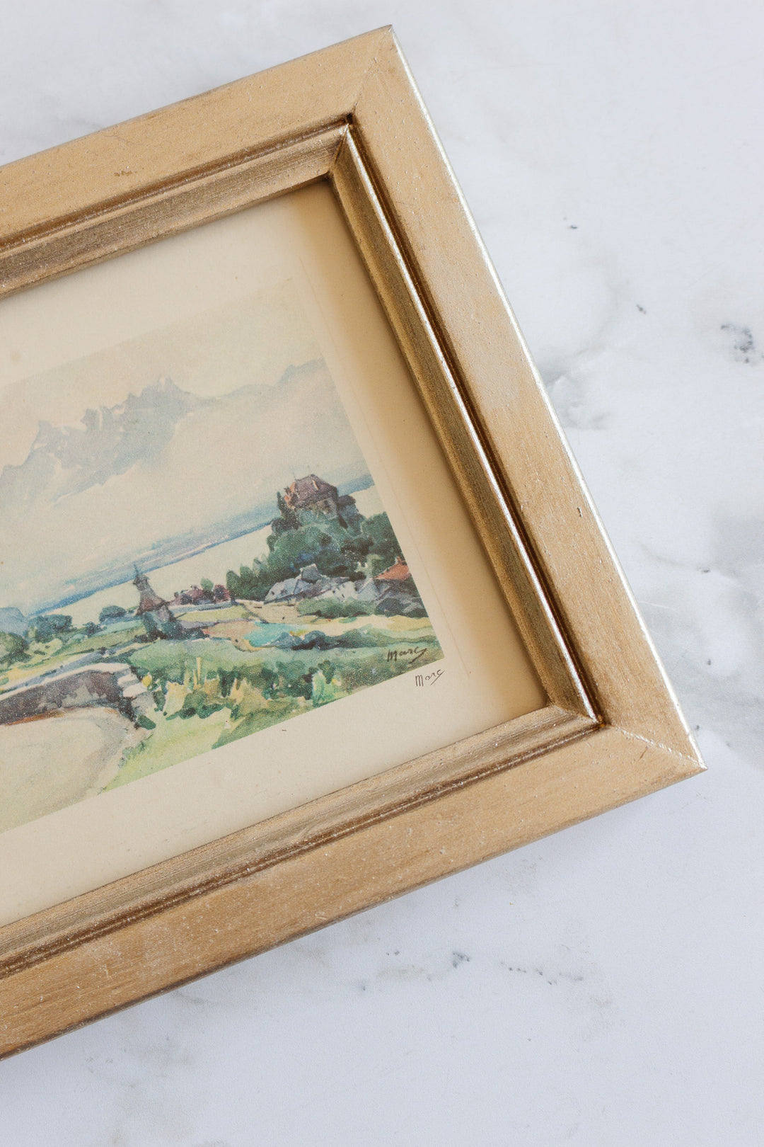 The art features a landscape watercolor painting in soft pastel colors, depicting trees, water, and buildings. It is set in a gold-finished wooden frame with elegant dimensions and rests on a white marble surface.