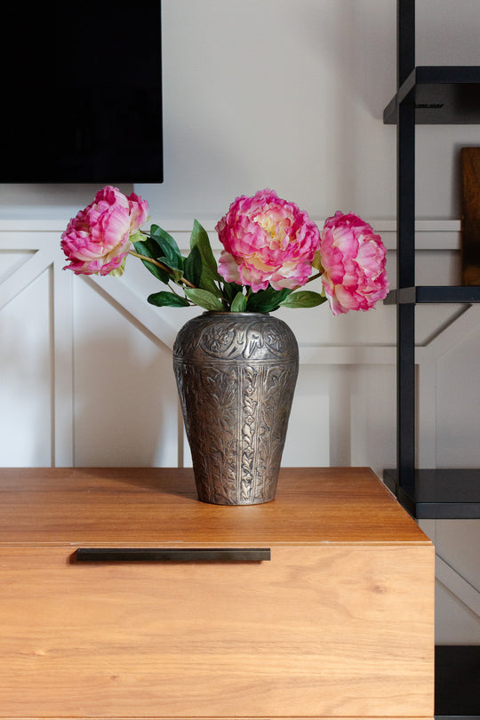 A metal vase holds three pink peonies with green leaves, displayed on a wooden surface near a black shelf against a white wall.