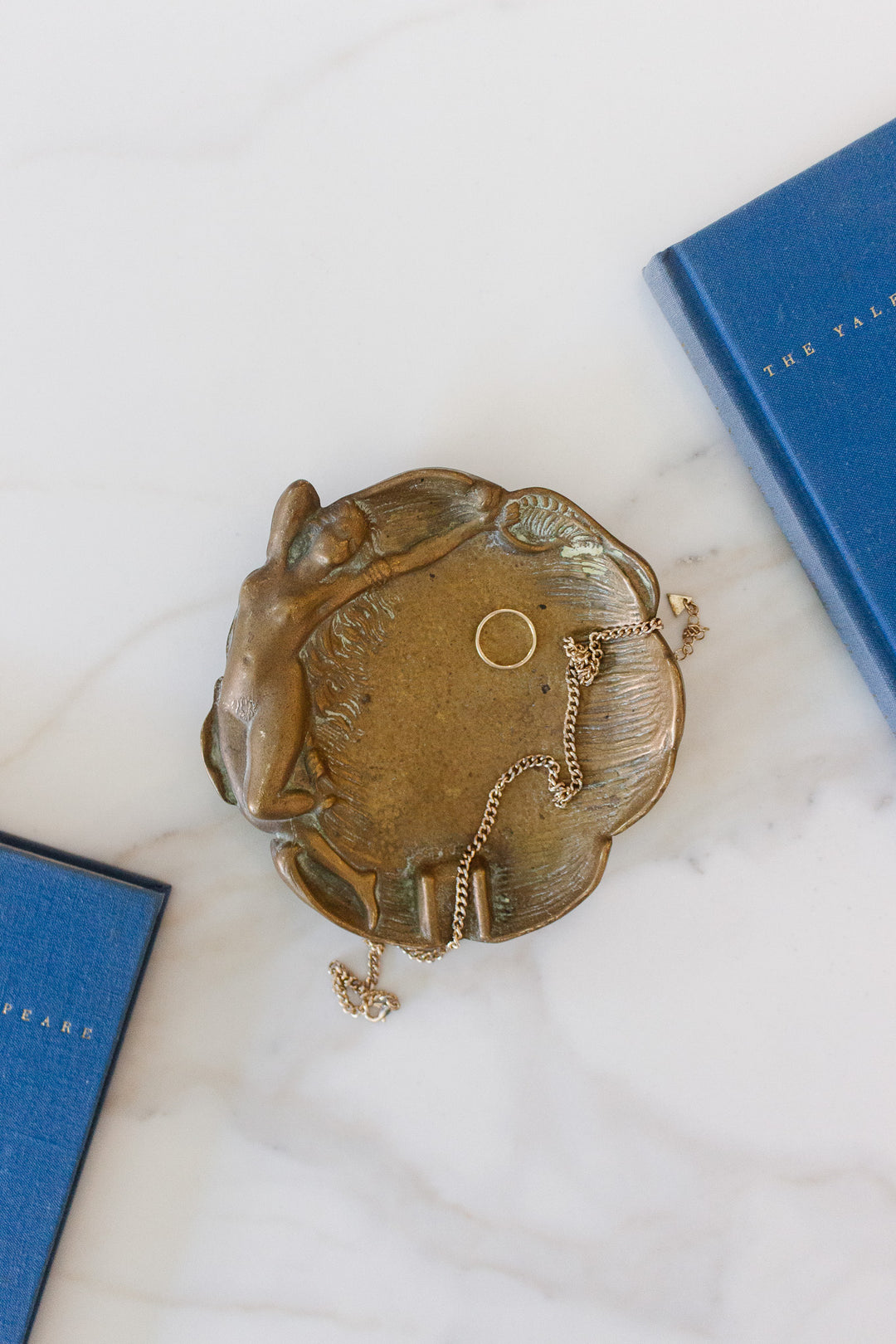 A brass trinket dish, elegantly sized, holds a gold ring and chain necklace on a white marble surface next to two blue hardcover books.