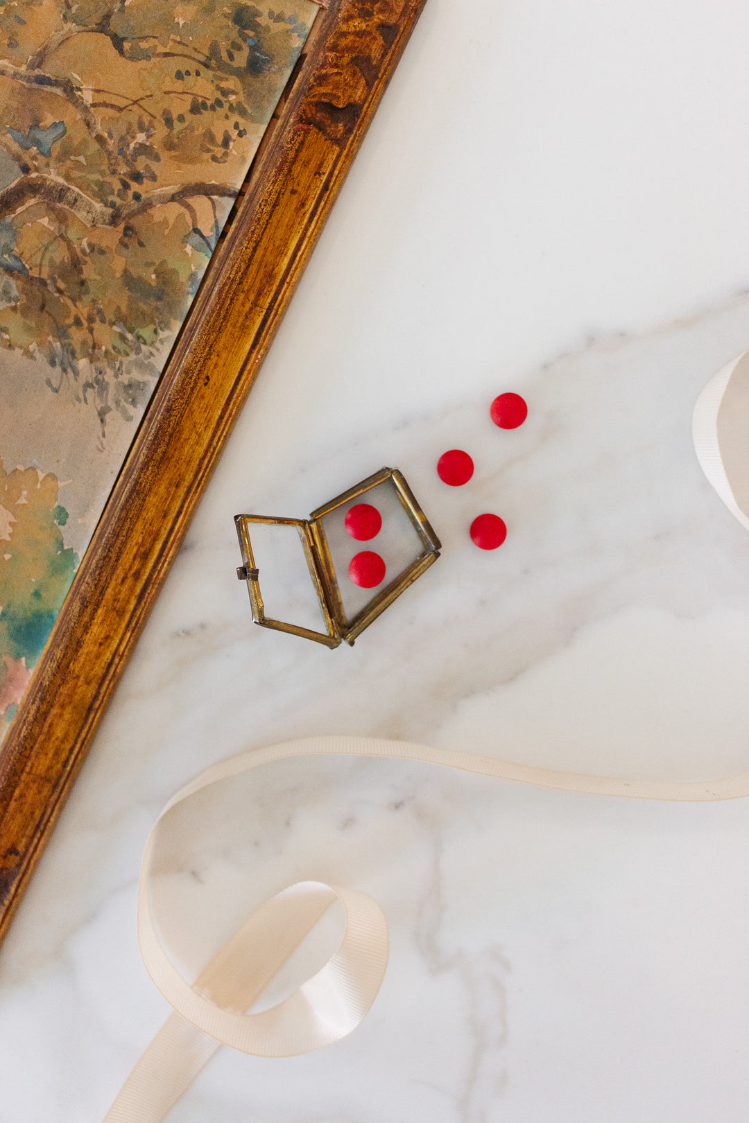 A small glass pill box sits open on a marble surface with five red round candies scattered nearby, accented by a cream-colored ribbon and a framed painting partially visible at the edges.