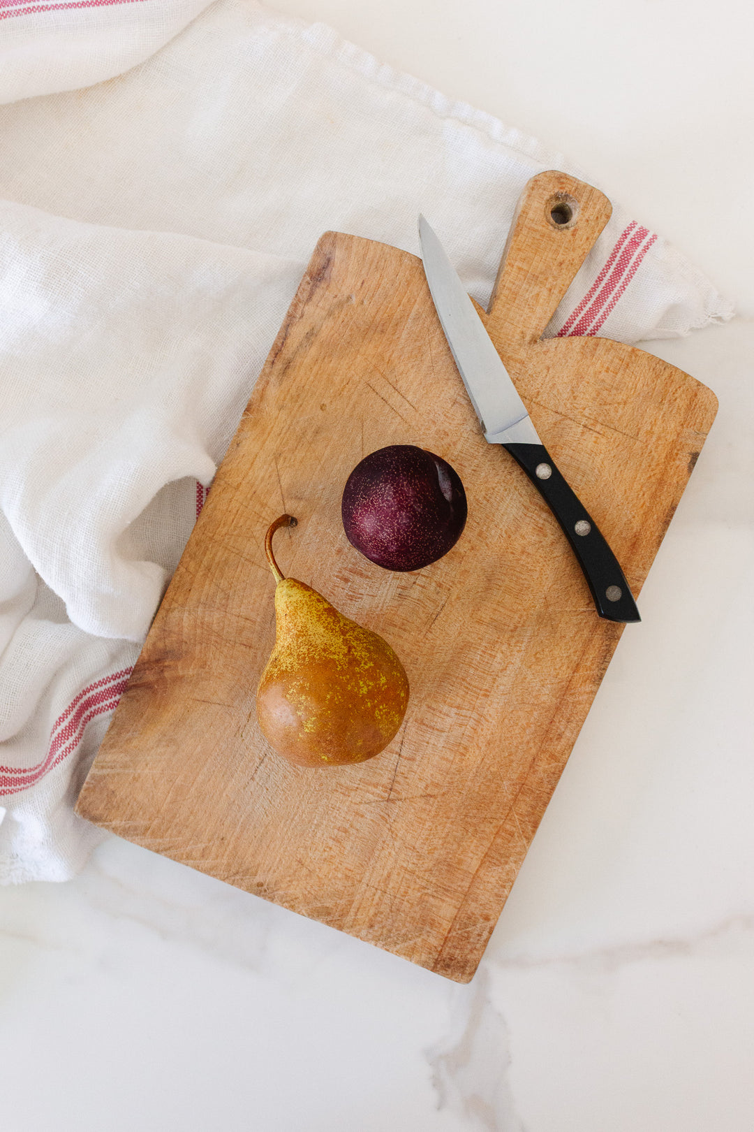 A breadboard with a knife, a brown pear, and a dark purple plum rests on a white surface beside a white cloth with red stripes, showcasing the breadboard’s clean lines and compact size.