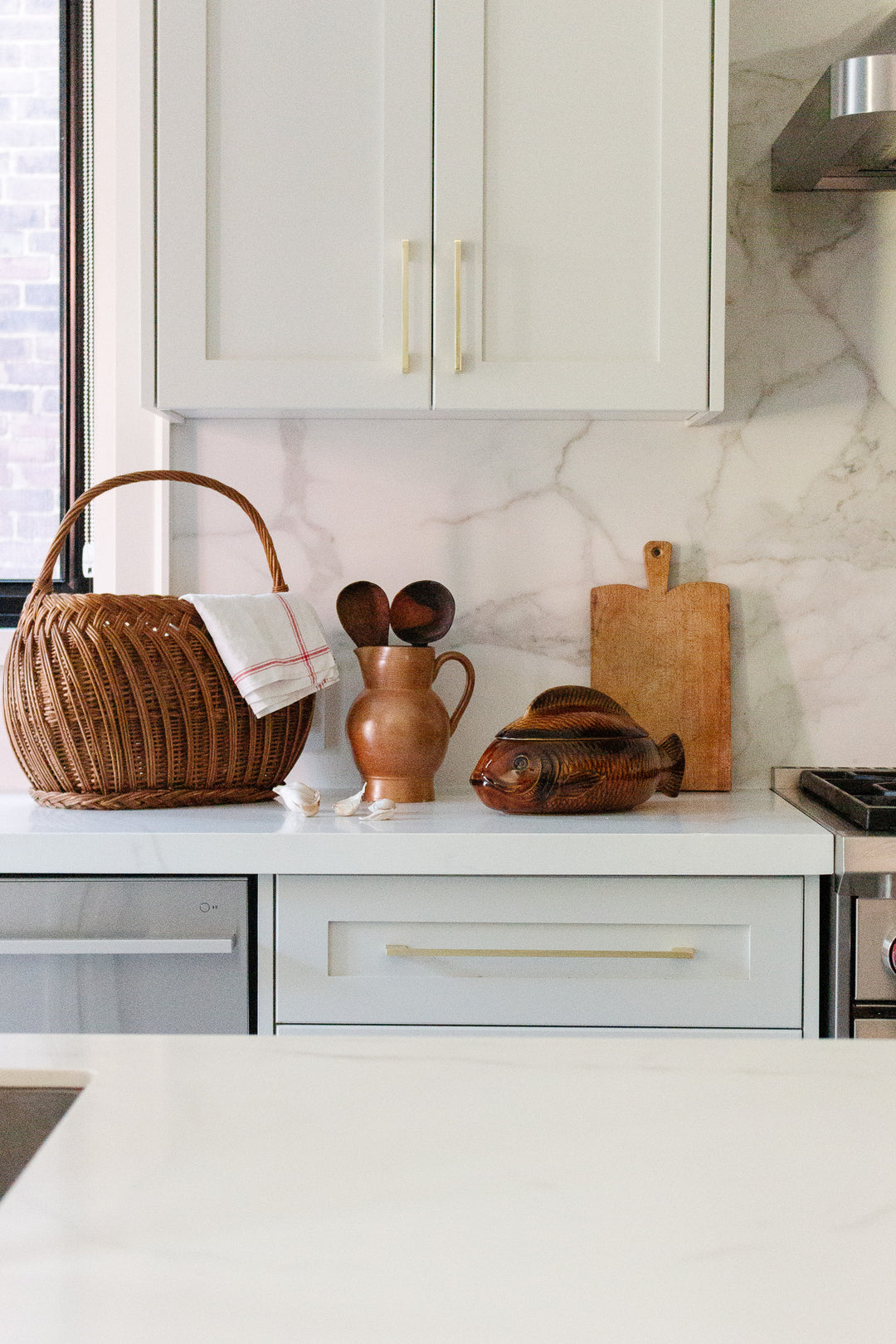 A kitchen counter featuring a wicker basket, brown ceramic jug with wooden utensils, the "fish soupiere" wooden container, small cutting board, and folded cloth set against a stylish white marble backsplash.