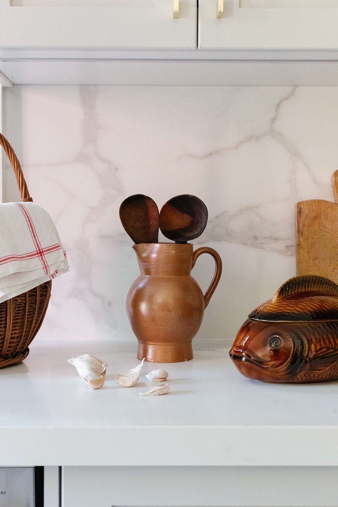 A kitchen counter displays a large jug holding wooden spoons, a wicker basket with a cloth, garlic cloves, a wooden fish sculpture, and a cutting board, all neatly arranged against a marble-pattern backsplash for maximum space.