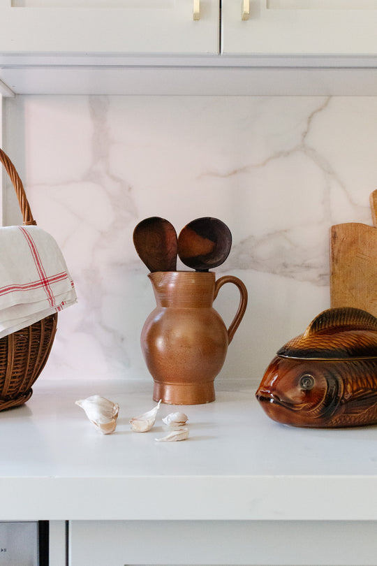 A kitchen counter displays a large jug holding wooden spoons, a wicker basket with a cloth, garlic cloves, a wooden fish sculpture, and a cutting board, all neatly arranged against a marble-pattern backsplash for maximum space.