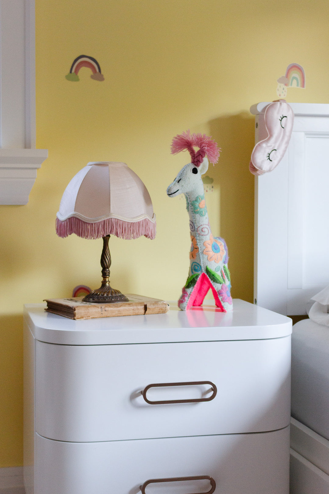 A compact white nightstand, ideal for small spaces, features a lamp beside a bed against a yellow wall decorated with rainbows and a hanging sleep mask. On the nightstand rest an old book, plush giraffe, and pink triangle.