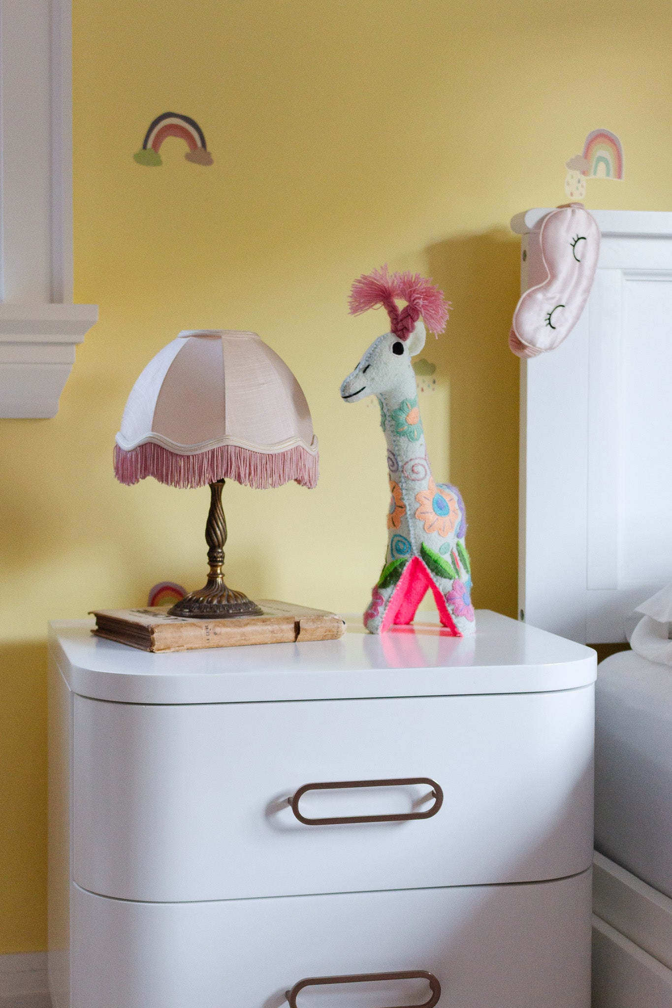 A compact white nightstand, ideal for small spaces, features a lamp beside a bed against a yellow wall decorated with rainbows and a hanging sleep mask. On the nightstand rest an old book, plush giraffe, and pink triangle.