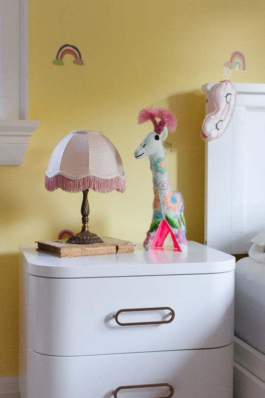 A compact white nightstand, ideal for small spaces, features a lamp beside a bed against a yellow wall decorated with rainbows and a hanging sleep mask. On the nightstand rest an old book, plush giraffe, and pink triangle.