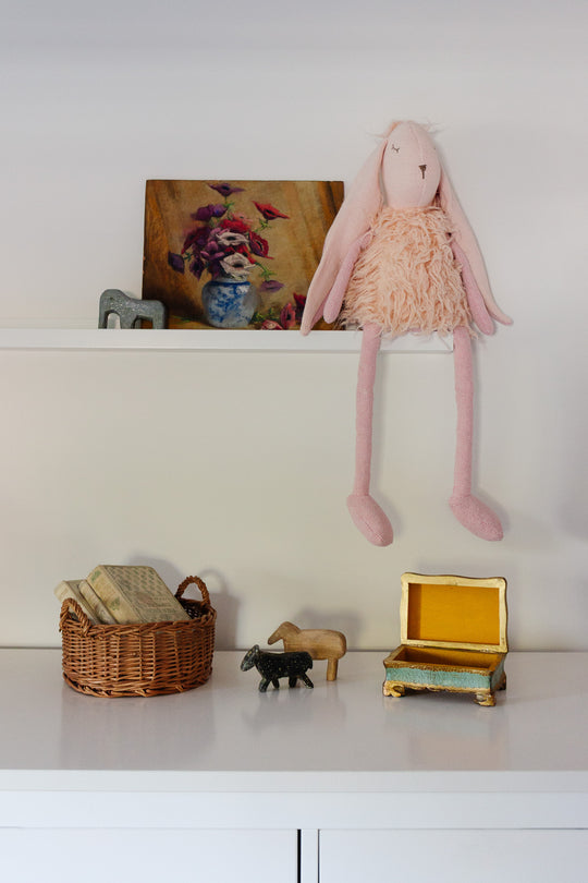 A shelf holds a pink stuffed bunny, a floral painting, and a decorative letter. Below, on a white surface, rests the basket filled with books along with two animal figurines and an open jewelry box of different sizes.