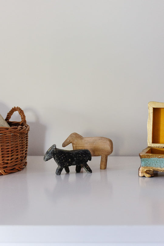 Two wooden animal figurines, one black and one natural, from the "wooden animals" collection are displayed on a white shelf. A woven basket is on the left, and a rustic yellow-and-blue box sits on the right against a plain wall.