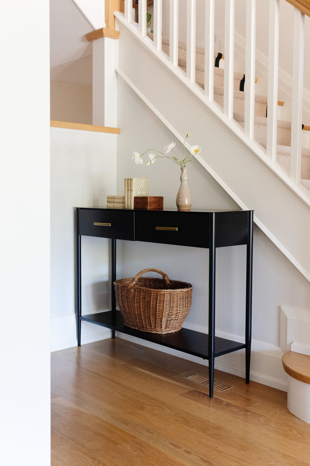 A wood box sits against the white-railed staircase on the wooden floor. On top, there are two books and a vase with white flowers. A wicker basket is placed on the lower shelf of this storage piece, which features two drawers.