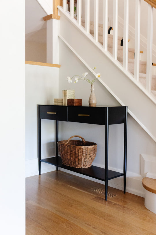A wood box sits against the white-railed staircase on the wooden floor. On top, there are two books and a vase with white flowers. A wicker basket is placed on the lower shelf of this storage piece, which features two drawers.