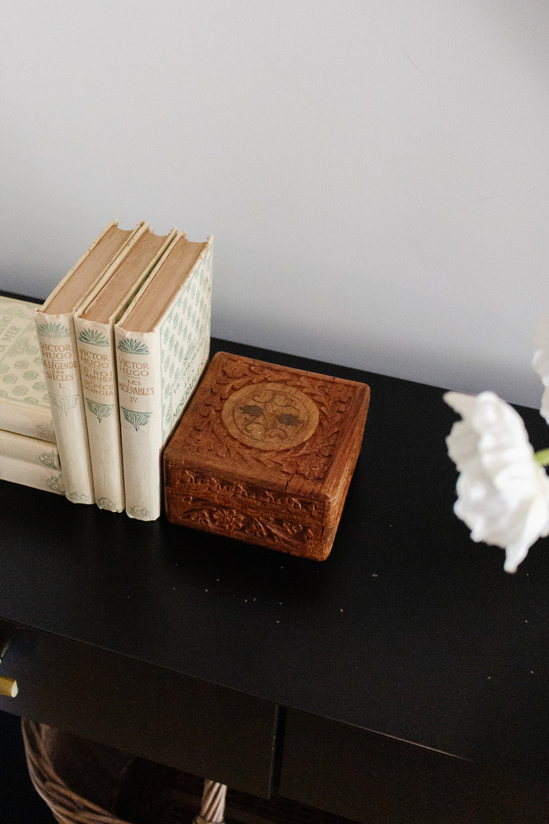 A wood box and four vintage books, each with unique dimensions, are neatly arranged on a black surface. White flowers are partially visible on the right side of the image.