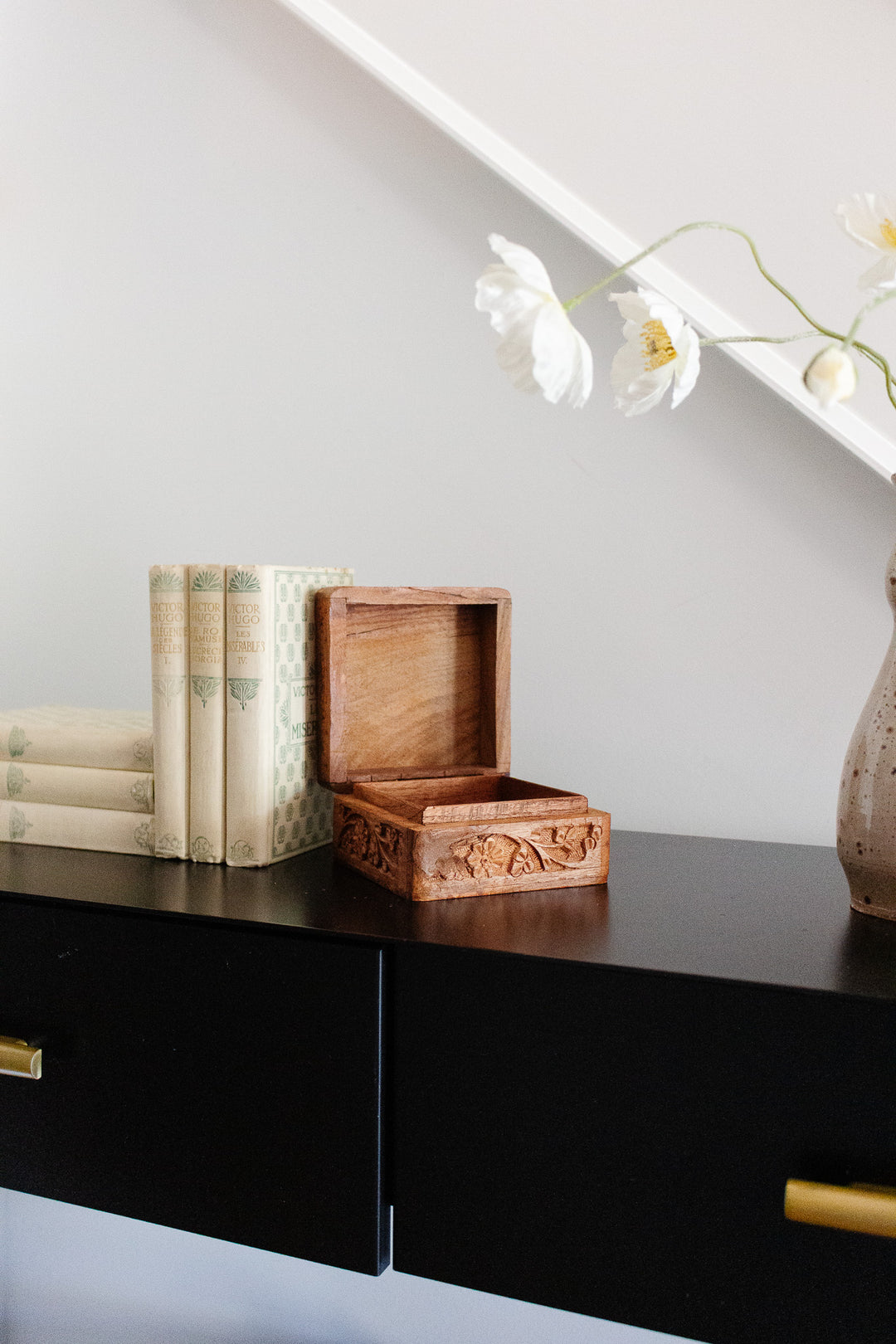 An open wood box with elegant dimensions sits on a black table next to vintage books, with a ceramic vase of white flowers in the background.