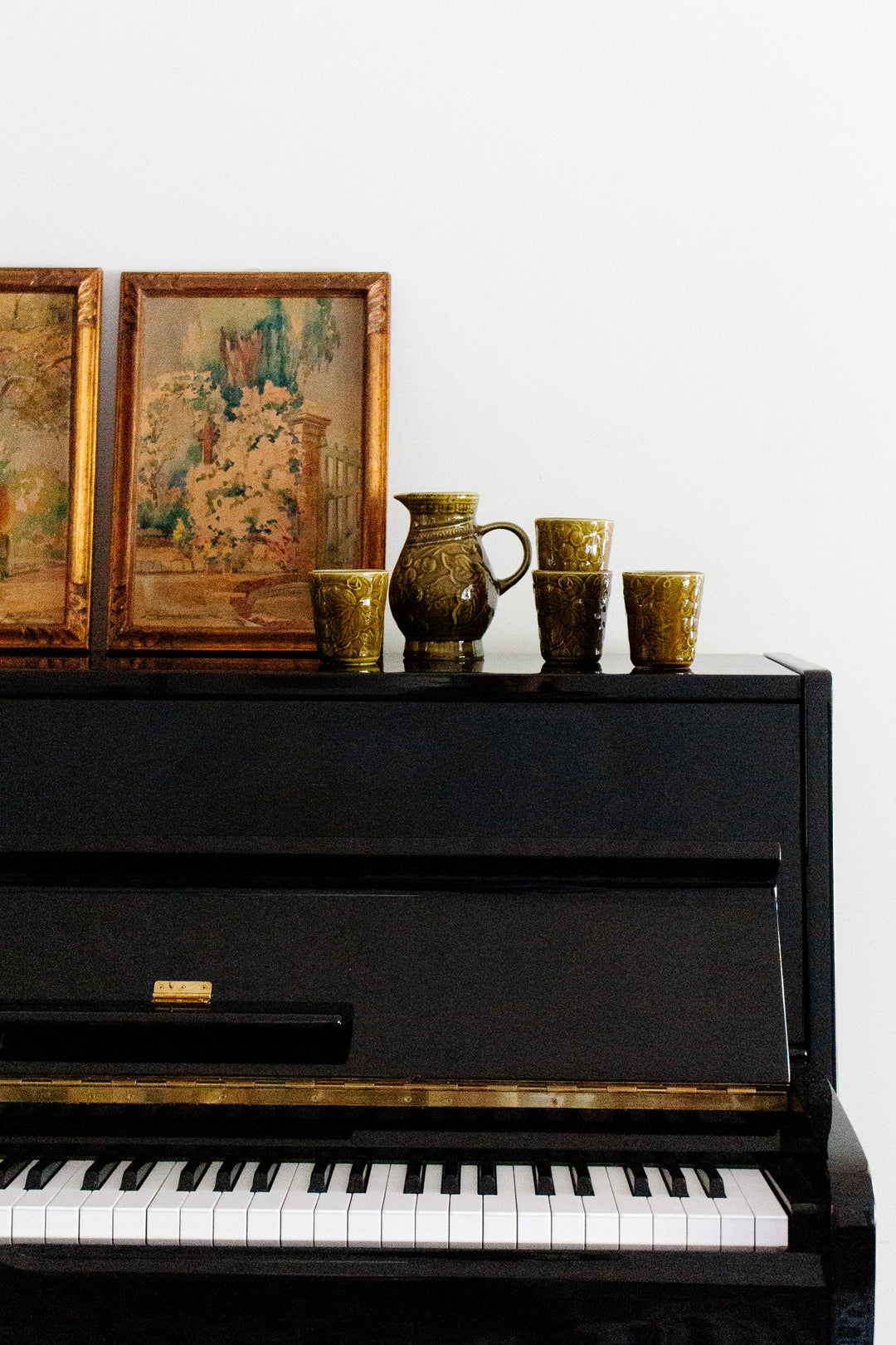 A black upright piano with visible keys is topped with two framed paintings and a drinkware set, featuring a jug and four cups in green ceramic, arranged against a plain white wall.