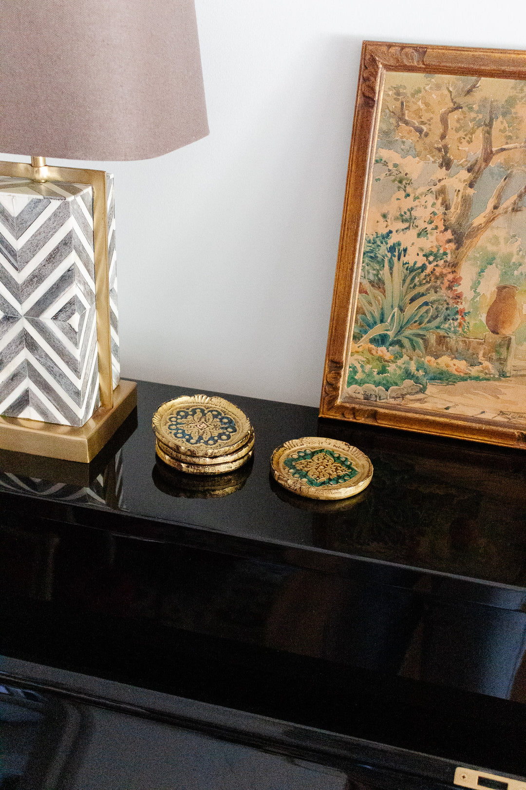 Florentine coasters, a decorative lamp, and a framed painting sit atop a black piano against a white wall, showcasing the balanced proportions of the arrangement.