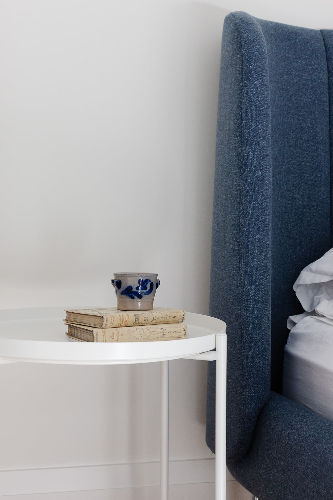 A small white side table with perfect dimensions holds a stack of three old books and a salt cellar, next to a blue upholstered bed with white sheets against a plain white wall.