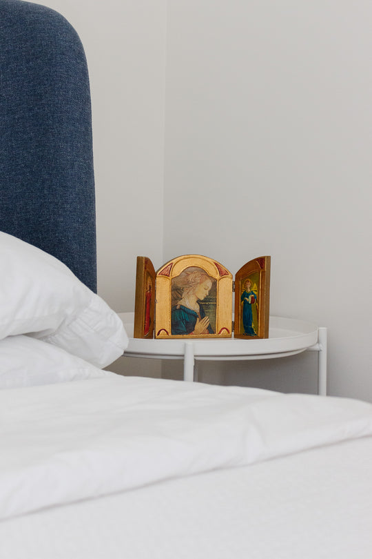 A "religious thing" with painted figures and a compact size rests on a white bedside table beside a bed with white linens and a blue upholstered headboard, creating a serene atmosphere in the light-walled room.