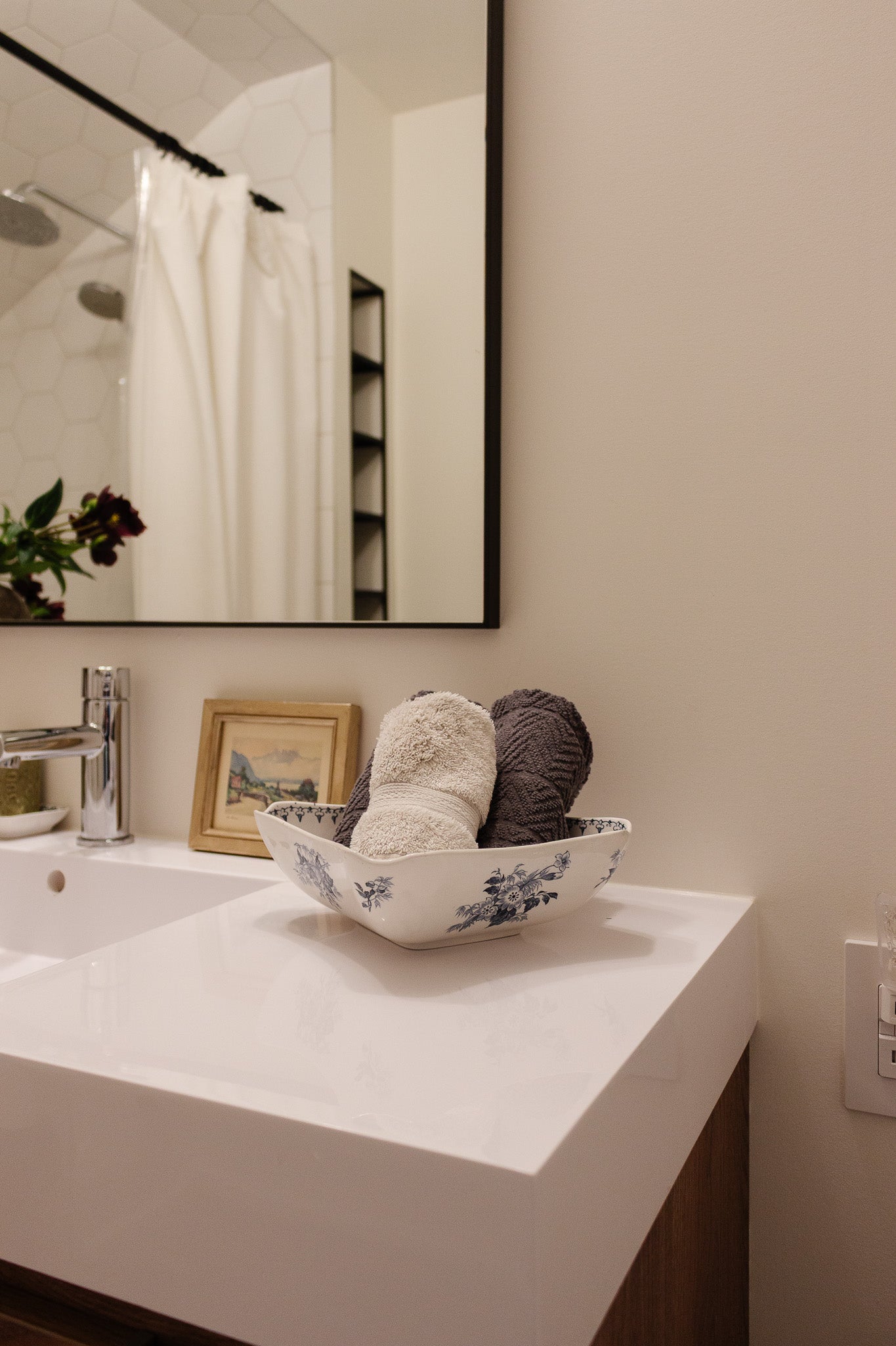 A modern bathroom countertop features a white sink, a floral bowl holding rolled towels, and décor accents. The mirror reflects the spacious shower with a white curtain.