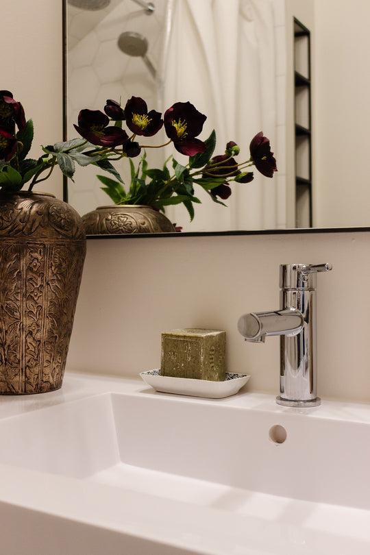 A modern bathroom sink with a chrome faucet, decorative vase of dark purple flowers, and a green bar of soap in a soap holder are reflected in the large mirror above, highlighting stylish details and balanced dimensions.