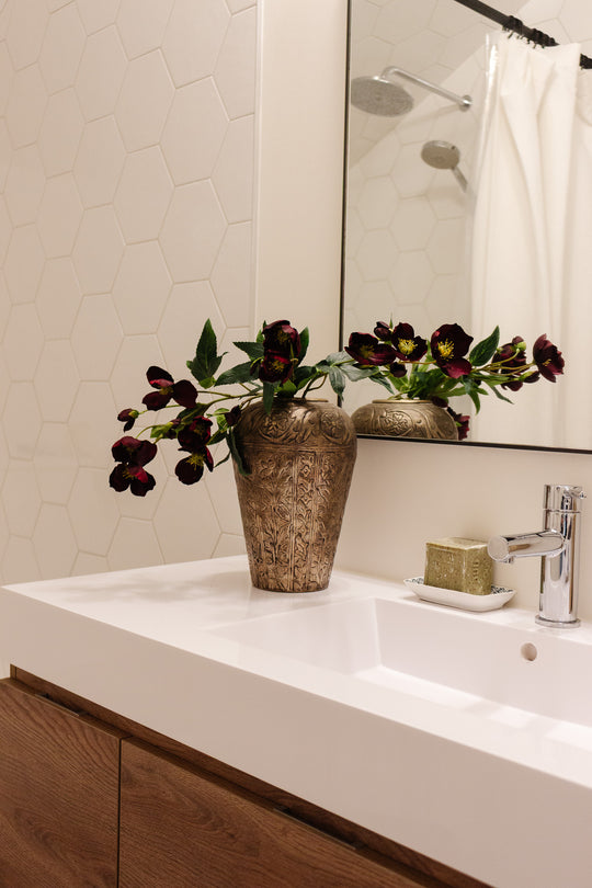 A metal vase with dark red flowers sits on a modern white bathroom sink beside a bar of soap and faucet, set against a hexagonal tile wall and mirror that reflects the elegant arrangement.