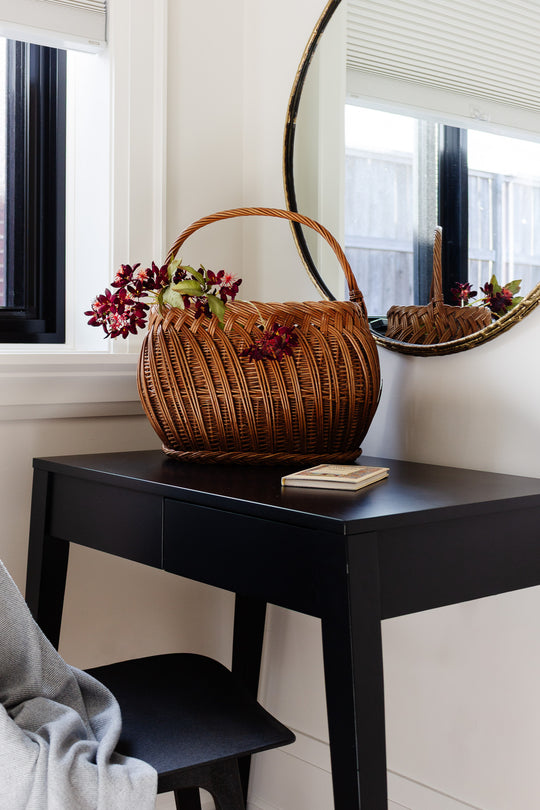 A basket sits on a black desk beneath a round mirror in a bright room near a window, with a closed book and black chair nearby.