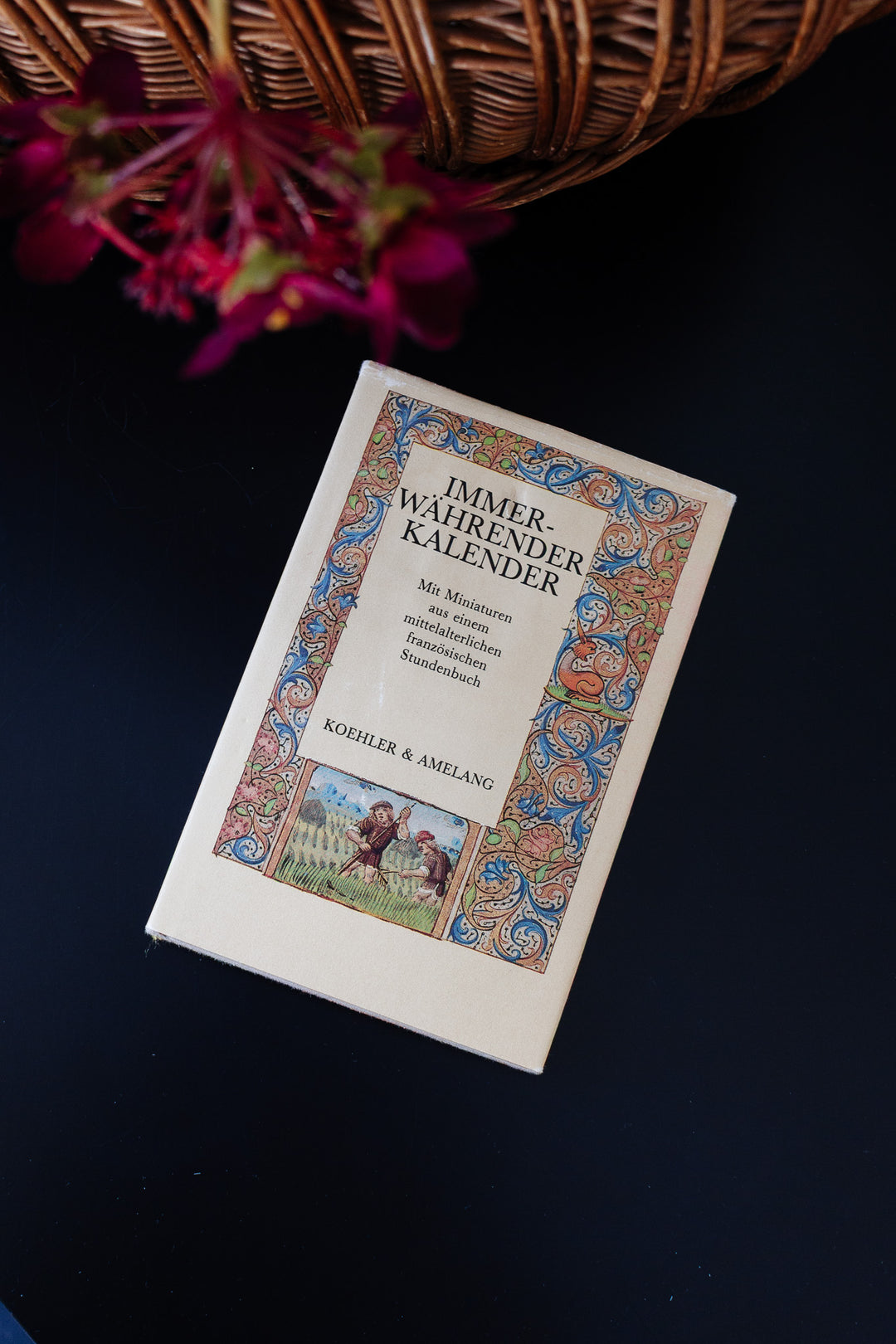 A book with an ornate, colorful border on the cover and striking dimensions from publisher Koehler & Amelang rests on a dark surface beside a wicker basket and reddish flowers.