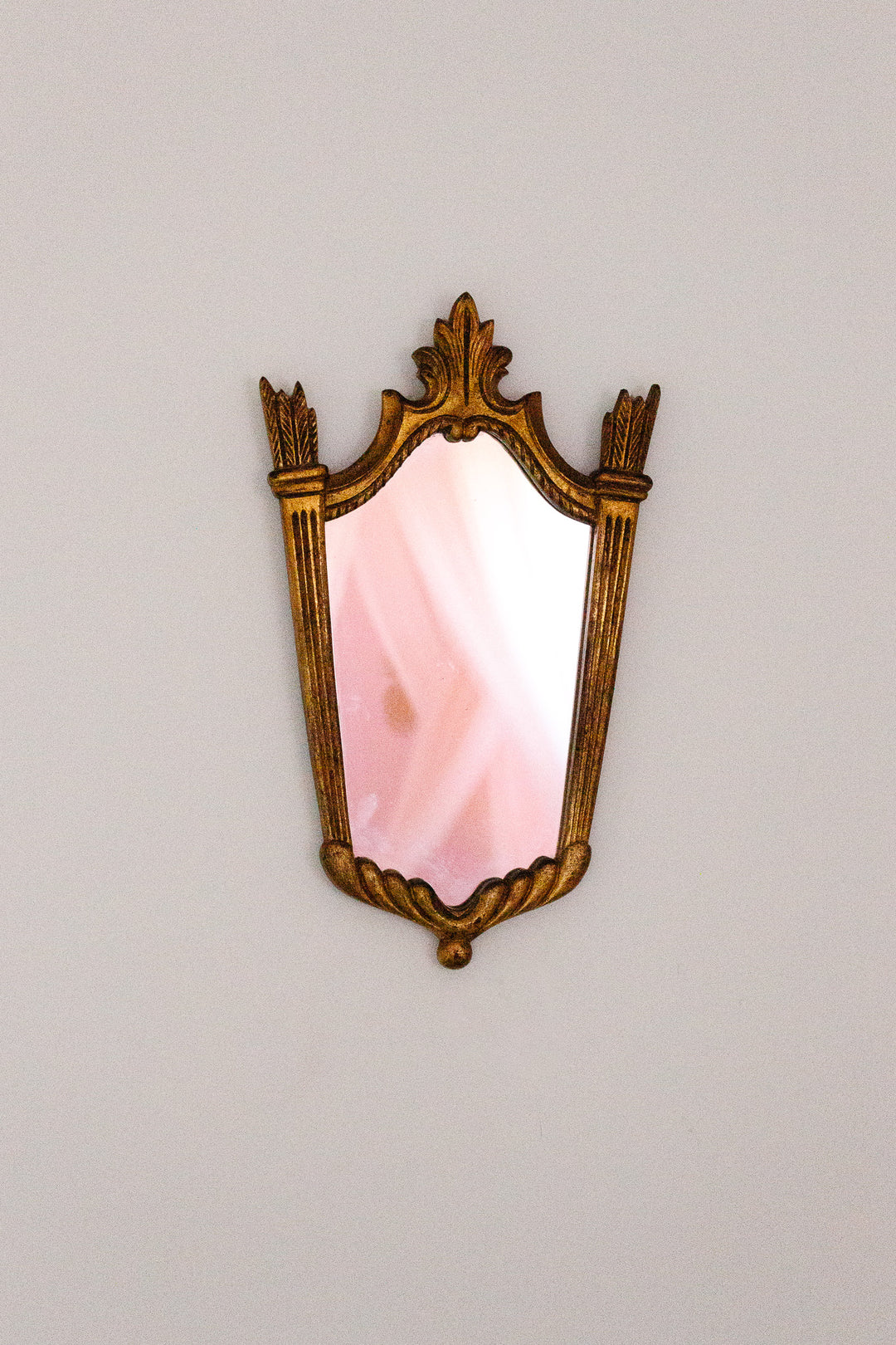A vintage-style mirror with a gold frame hangs on a white wall. The mirror reflects soft pink and white tones, possibly from a room or curtain, and adds an elegant touch with its ornate design.