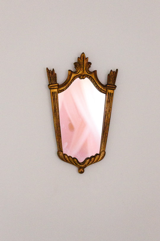 A vintage-style mirror with a gold frame hangs on a white wall. The mirror reflects soft pink and white tones, possibly from a room or curtain, and adds an elegant touch with its ornate design.