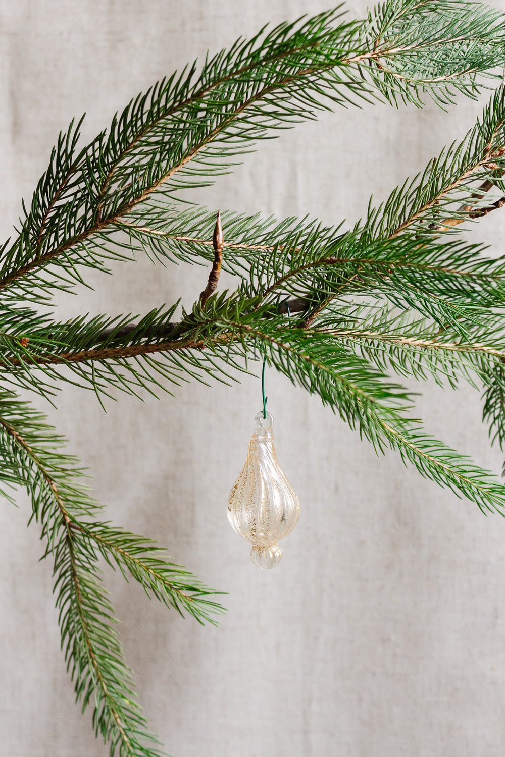 An antique Italian hand-blown glass ornament with aventurine gold inclusions hangs from a green pine branch, set against a neutral beige background.
