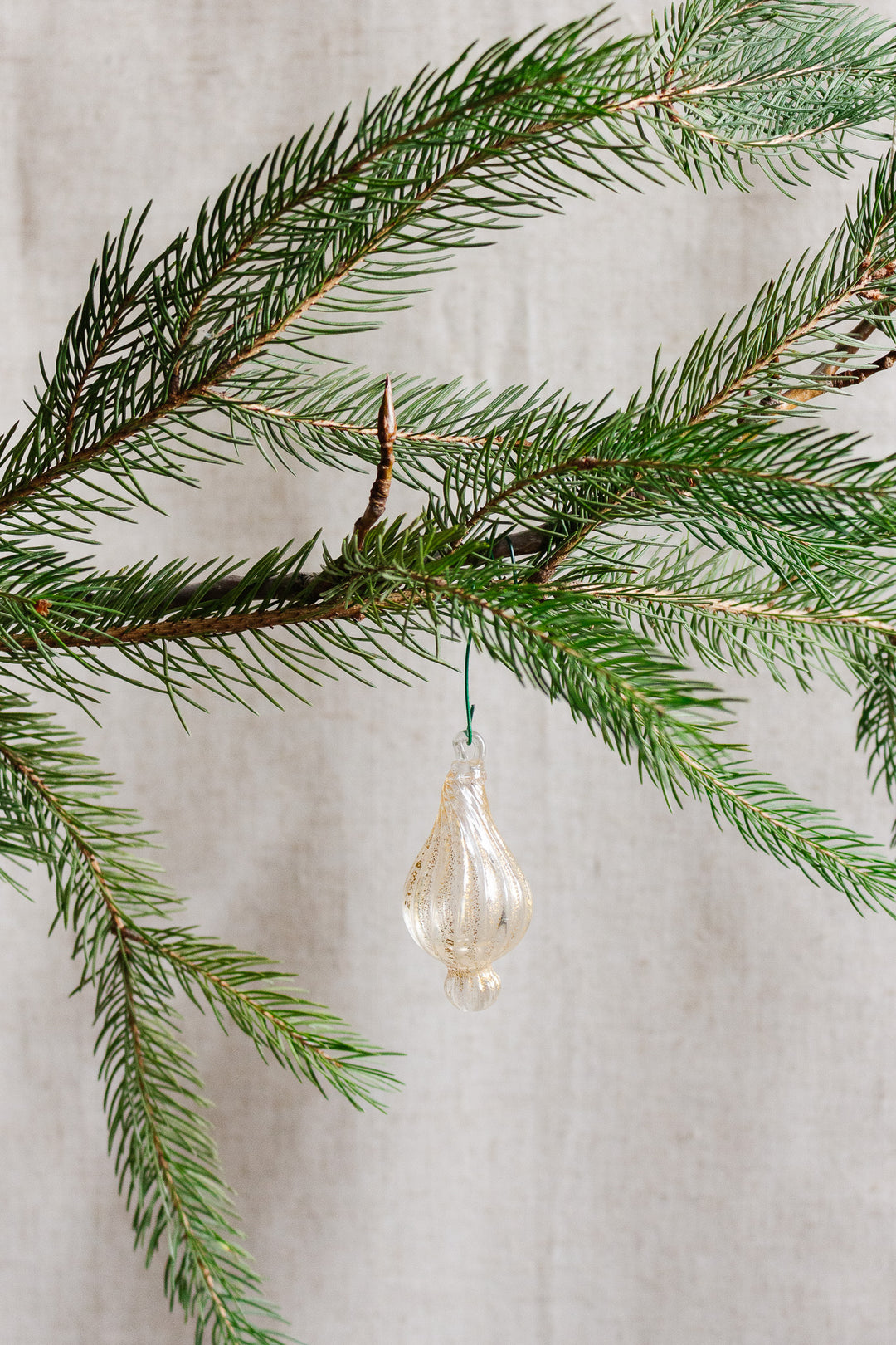 An antique Italian hand-blown glass ornament with aventurine gold inclusions hangs from a green pine branch, set against a neutral beige background.