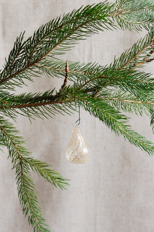 An antique Italian hand blown glass ornament with aventurine gold inclusions, shaped like a teardrop, hangs from a green pine branch against a textured beige background.