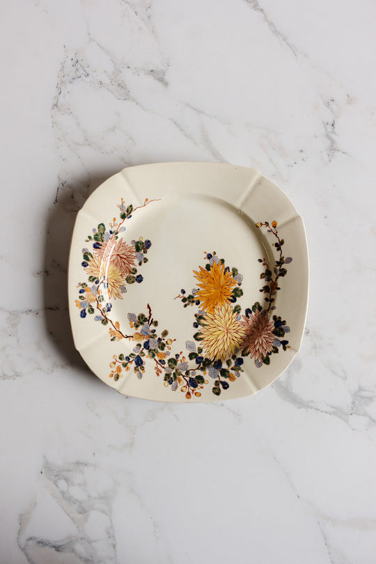 A turn of the century Gien transferware cake plate in cream, square-shaped with rounded edges, features yellow and pink flowers with green leaves along one side and sits elegantly on a white marble surface.