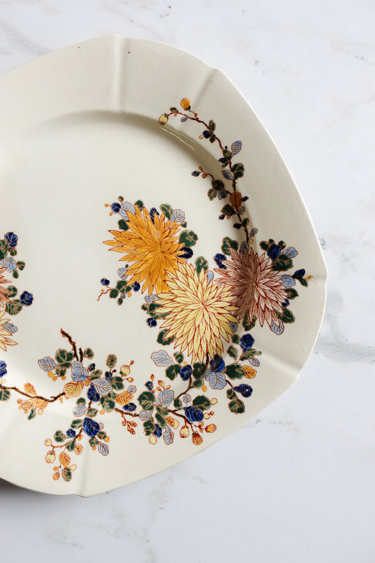 A turn of the century Gien transferware cake plate with a vintage cream base, scalloped edges, and yellow, orange, and blue floral motifs rests on white marble—an elegant accent for any French-inspired setting.