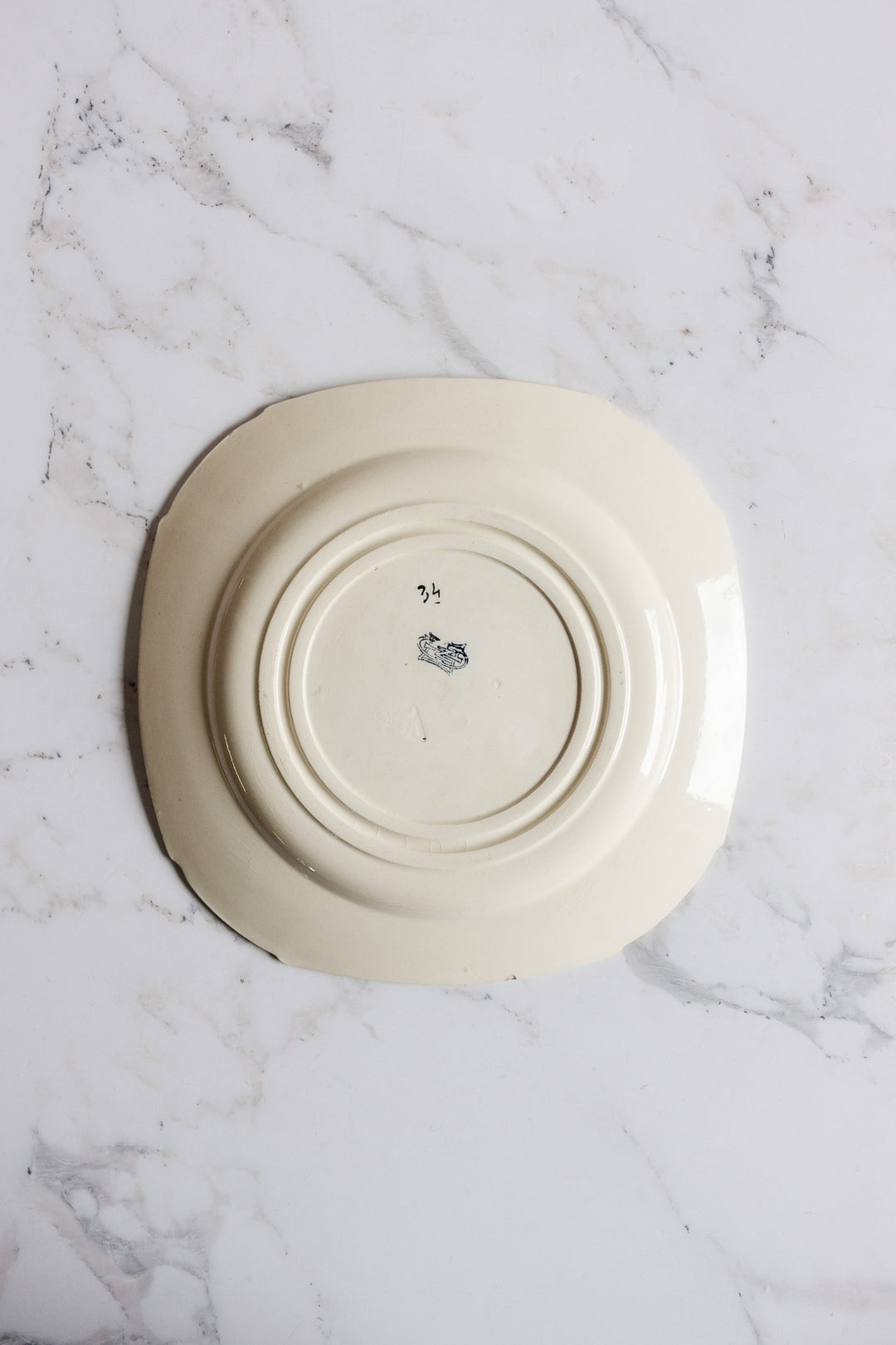 A turn of the century Gien transferware cake plate, cream-colored and square, rests upside down on white marble, revealing its base with a small central marking.
