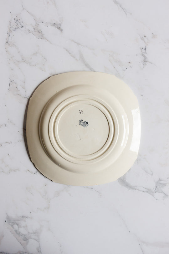 A turn of the century Gien transferware cake plate, cream-colored and square, rests upside down on white marble, revealing its base with a small central marking.
