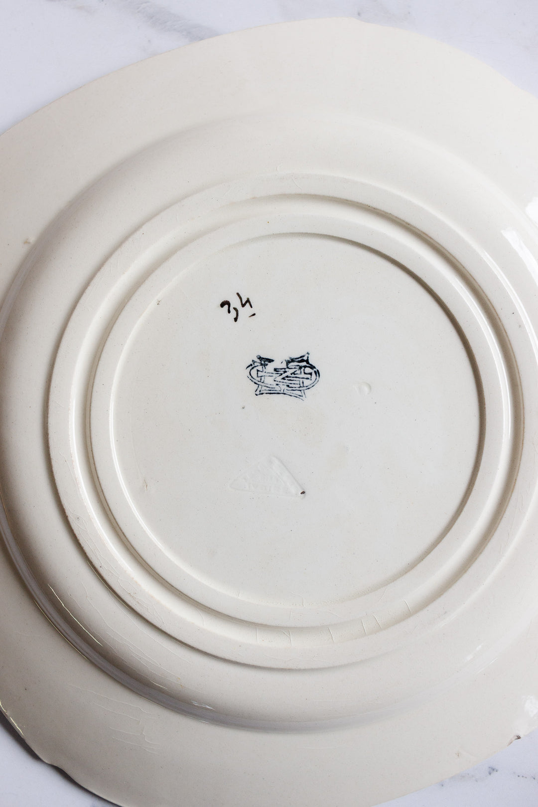The underside of this turn of the century Gien transferware cake plate features a hand-drawn 34 in black ink, a dark decorative stamp or logo, and visible circular ridges, all resting elegantly on a light French table.