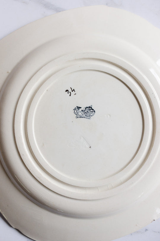 The underside of this turn of the century Gien transferware cake plate features a hand-drawn 34 in black ink, a dark decorative stamp or logo, and visible circular ridges, all resting elegantly on a light French table.