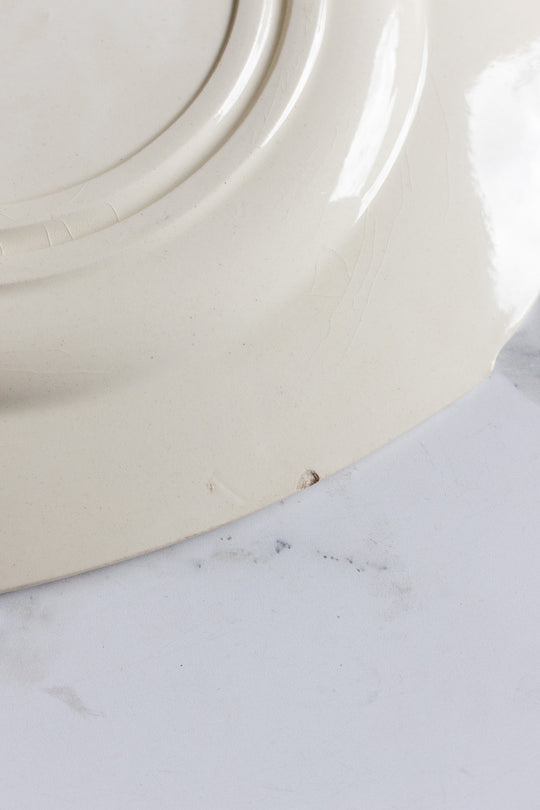 A close-up of the underside of a turn of the century Gien transferware cake plate, off-white in color with a small chip on the rim, resting on a white marble surface.