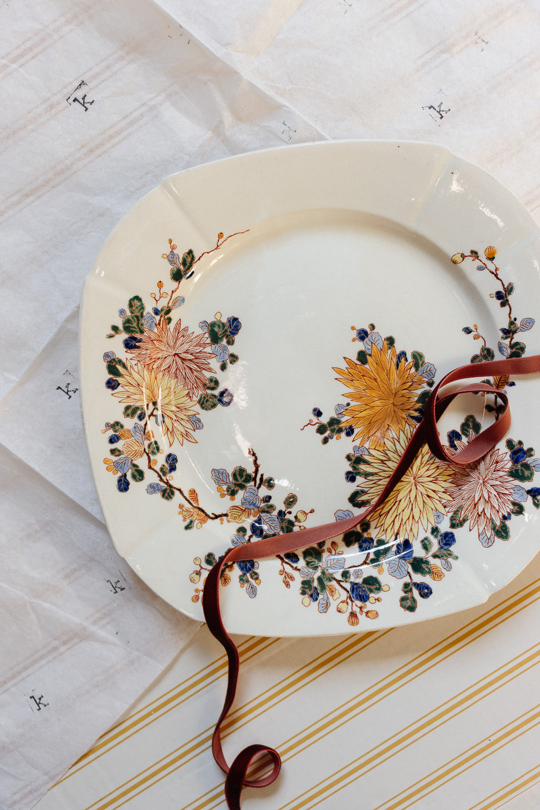 A turn of the century Gien transferware cake plate with yellow and blue floral designs rests on striped, lettered paper, accented by a brown ribbon for a charming French-inspired table setting.