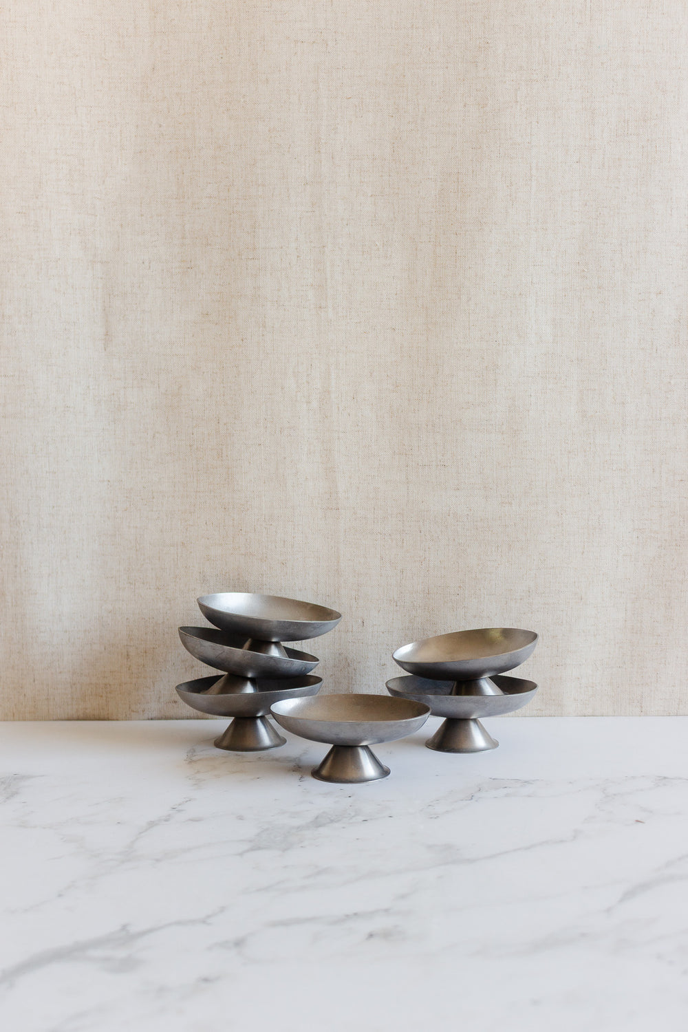 Six vintage French stainless steel oblong ice cream cups with pedestal bases are stacked on a marble surface, set against a beige fabric background.