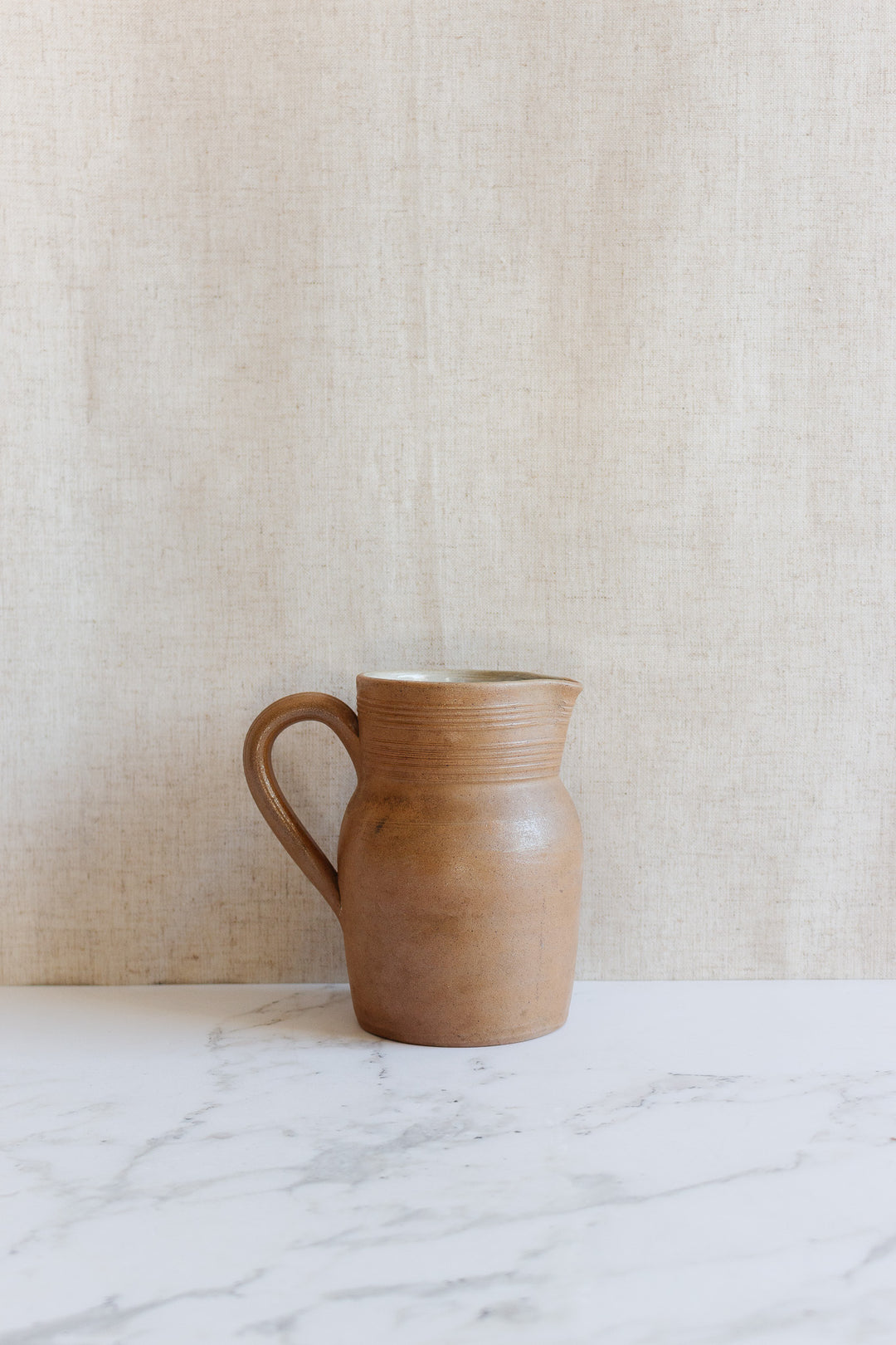A vintage French "grès de berry" stoneware pitcher with a handle sits on a white marble surface against a beige textured background.